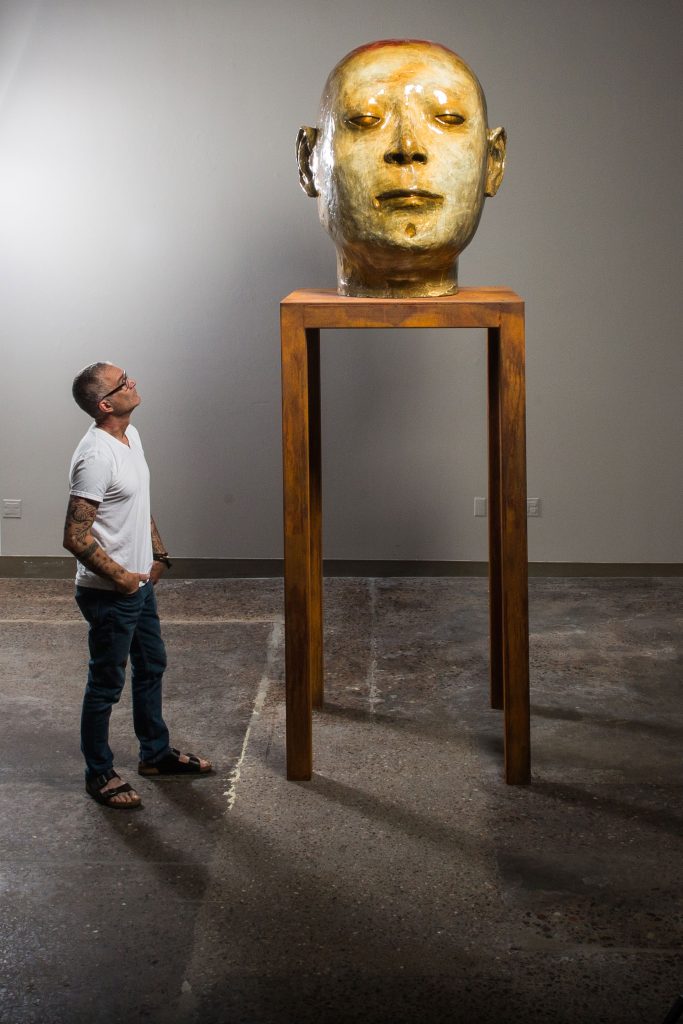 Artist looking up at a sculpture of a head on a tall pedastal