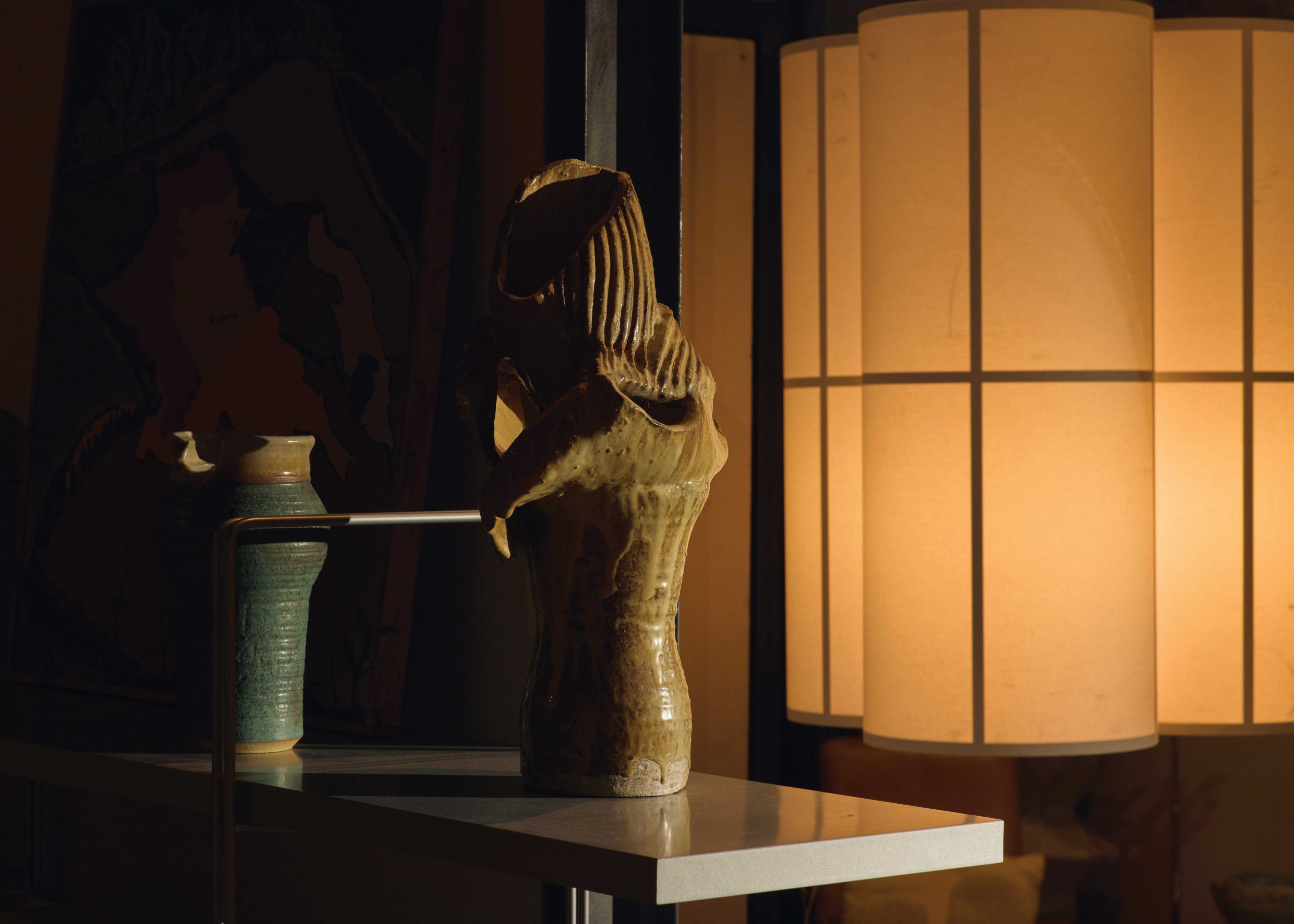 A hanging pendant light provides subtle illumination for ceramic works by Philip Bellomo displayed on an adjacent shelf.