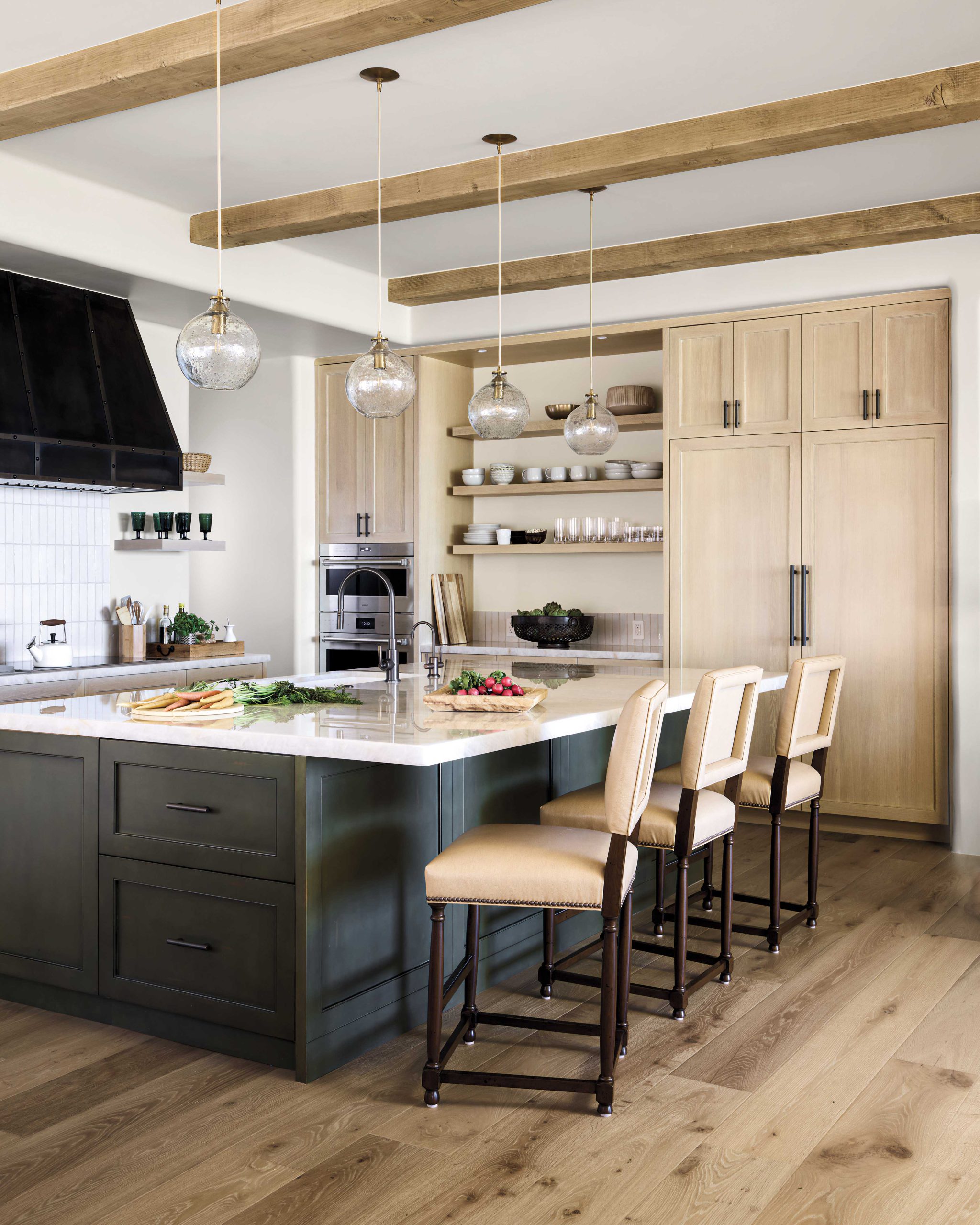 The relocated kitchen features pale, distressed wood cabinetry contrasted by the island’s deep stone-hued tones. Barstools are transitional, in keeping with the furnishings theme of the interior.