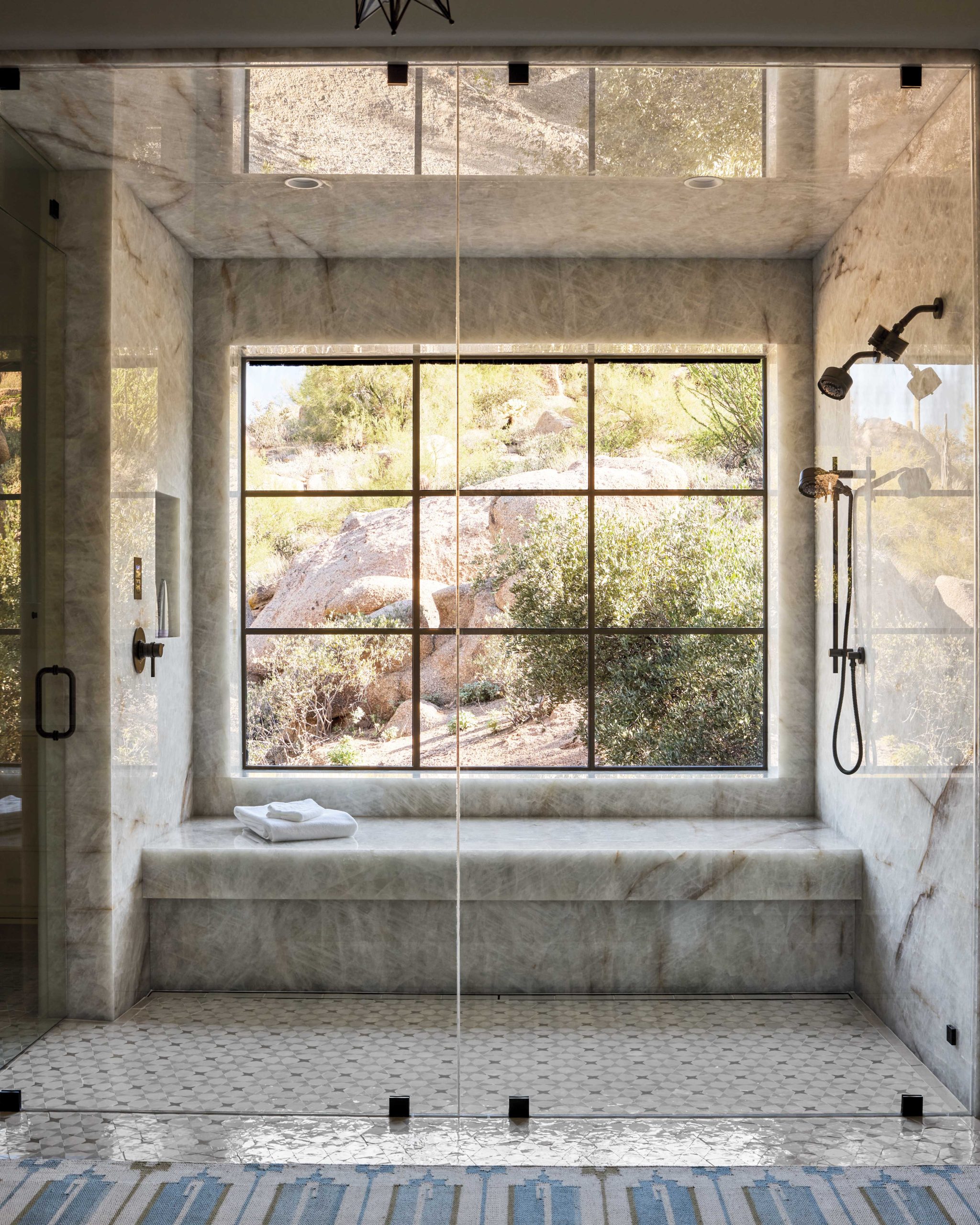 Several smaller rooms were eliminated to create a more spacious primary suite. In the bathroom, the steam shower’s window faces a private hillside, while the vanity cabinetry’s patterning is a nod to vintage Southwest. The bedroom’s serene tones allow the framed outdoor view to be the centerpiece.